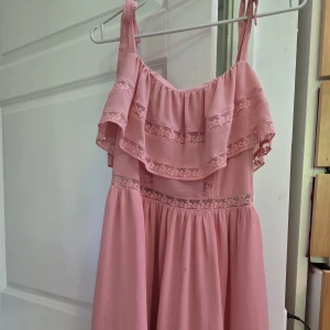 Rosa kortklänning med volang och spets - Supersöt rosa kortklänning med volang upptill och fina spetsdetaljer. Klänningen har smala axelband med knyt och ett luftigt, flowy tyg som känns perfekt för varma dagar.