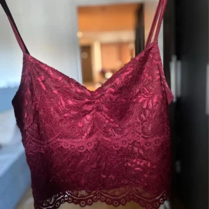 Vinröd spetsig linnetopp - Supersnygg vinröd linnetopp i spets med tunna axelband och v-ringning. Croppad modell med blommigt spetsmönster och vågig kant nertill. Perfekt för att lyfta vilken outfit som helst med en romantisk touch.