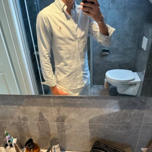 Randig skjorta från Ralph Lauren S - Säljer en klassisk slim fit skjorta från Ralph Lauren i storlek S. Skjortan är vit med tunna blå ränder, har lång ärm, knappar framtill och den ikoniska broderade loggan på bröstet. Tillverkad i mjuk bomull. Dock saknar skjortan en knapp (se sista bild)