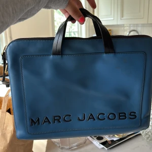 Datorväska från Marc Jacobs - Snygg blå datorväska från Marc Jacobs i slät skinn med svarta handtag och stor logga framtill. Väskan har dragkedja upptill och är perfekt för laptop eller dokument. Enkel, stilren design som sticker ut med sin djupa blå färg och guldiga detaljer på dragkedjan.