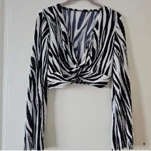 Zebramönstrad croppad topp från SHEIN - Säljer en croppad långärmad topp från SHEIN med svartvitt zebramönster. Toppen har omlott framtill med twistad detalj och är tillverkad i lätt, plisserad polyester. Perfekt för dig som vill sticka ut med en edgy vibe.