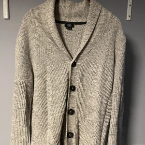 Grå kofta från F&F med knappar - Säljer en grå stickad kofta från F&F i storlek M. Koftan har bred krage, långa ärmar och stängs med fyra svarta knappar framtill. Den är ribbstickad på ärmar och nedre delen, vilket ger en snygg struktur. Perfekt för lager-på-lager under kyliga dagar.