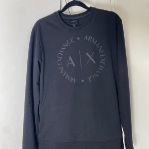 Svart sweatshirt från Armani Exchange - Svart sweatshirt från Armani Exchange med rund hals och stort A|X-logotryck framtill. Tröjan har lång ärm och en klassisk, avslappnad passform. Perfekt för dig som gillar stilrena och enkla plagg med diskret branding.