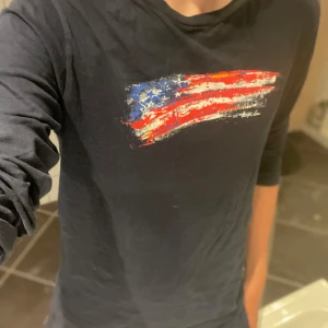 Mörkblå långärmad T-shirt Polo Ralph Lauren - Snygg mörkblå långärmad tröja från Polo Ralph Lauren med ett stort tryck av en amerikansk flagga i rött, vitt och blått på bröstet. Tröjan är i mjuk bomull och har en avslappnad passform, perfekt till jeans eller shorts.