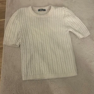 Vit kabelstickad tröja Gina Tricot - Kabelstickad vit tröja från Gina Tricot med korta ärmar och rund halsringning. Tröjan har en klassisk, ribbad kant vid ärmslut och nederkant. Perfekt för lager-på-lager och enkel att styla till olika looks.
