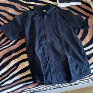 Svart kortärmad skjorta med ficka - En stilren svart skjorta med korta ärmar och klassisk krage. Skjortan har knäppning framtill och en bröstficka på vänster sida. Perfekt för dig som gillar enkel och clean stil. Materialet känns mjukt och lätt, troligen bomull eller polyester.