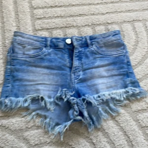 Blå jeansshorts med fransig kant - Säljer ett par blå jeansshorts med snyggt fransig kant och klassisk femficksmodell. Shortsen har en ljus tvätt och råa kanter som ger en cool, avslappnad vibe. Perfekta för varma dagar och festivalhäng.