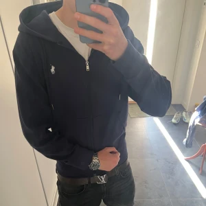 Mörkblå zip hoodie från Polo Ralph Lauren - Mörkblå hoodie med dragkedja från Polo Ralph Lauren storlek S-M. Klassisk design med huva, snörning och två fickor framtill. Ikoniska vita loggan broderad på bröstet. Skön bomullskvalitet och normal passform, perfekt för chill dagar.