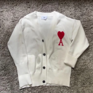 Vit kofta från Ami Paris med rött hjärta - Vit kofta från Ami Paris med svart knappstängning och rött hjärta med bokstaven A broderat på bröstet. Tjock ribbad mudd vid ärmslut och nederkant. Perfekt för lager-på-lager och stilrena outfits. PRIS EJ HUGGET I STEN!