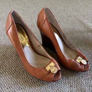 Bruna wedgepumps från Michael Kors  - Snygga bruna pumps med kilklack från Michael Kors i skinn. Skorna har öppen tå och dekoreras med guldfärgad MK-detalj framtill. Perfekta för dig som vill ha både höjd och komfort i en klassisk stil.