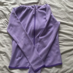 Lila träningsjacka med huva Kappahl - Sportig lila träningsjacka från Kappahl med huva och hel dragkedja framtill. Jackan är i mjuk polyester, har långa ärmar och diskreta fickor i sidorna. Perfekt för aktiva dagar och snygg att matcha med träningskläder.