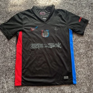 FC Barcelona Lamine Yamal tröja Nike - Säljer en svart FC Barcelona fotbollströja från Nike med Lamine Yamal och nummer 19 på ryggen. Tröjan har röda och blå sidopaneler, klubbmärke på bröstet och tryck i rött och grått. Tillverkad i lätt och ventilerande material, perfekt för match eller träning.