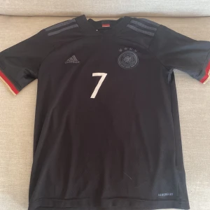 Tysklands Havertz #7 Adidas fotbollströja - Snygg svart Tyskland fotbollströja från Adidas med nummer 7 och namnet Havertz på ryggen. Tröjan har korta ärmar, diskreta grå ränder på axlarna och tysk flaggdetalj i nacken. Tillverkad i lätt och ventilerande Aeroready-material.