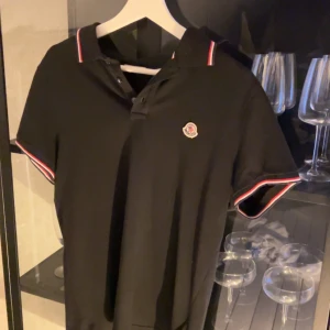 Svart Moncler pikétröja med röd/vit kant - Snygg svart pikétröja från Moncler med klassisk krage och knäppning framtill. Tröjan har Moncler-logga på bröstet och detaljer i rött och vitt på krage och ärmslut. Perfekt för dig som gillar stilrena och exklusiva plagg. OBS! Pris kan diskuteras!