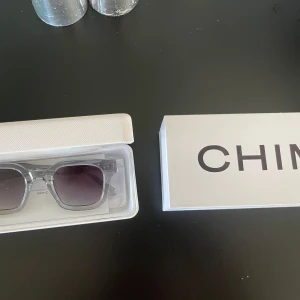 CHIMI transparenta solglasögon silver - Snygga solglasögon från CHIMI med transparent fyrkantig båge och silvriga glittriga detaljer på skalmarna. Glasen är mörktonade och ger en cool vibe. Kommer med allting man får med när man köper de!