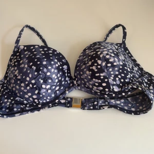 Bikinitopp - Oanvänd mörkblå bikinitopp från Röhnisch med vita prickar och justerbara axelband. Vadderade kupor. Metalknäppning baktill med logga på. Stl. S. Köparen står för fraktkostnad.