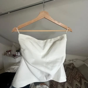 Vit axelbandslös tubtopp från Zara - Säljer en stilren vit tubtopp från Zara med axelbandslös bandeau-modell. Toppen har en lätt draperad framsida och är sydd i ett slätt tyg som ger en snygg siluett. Perfekt att matcha med jeans eller kjol för en clean look.