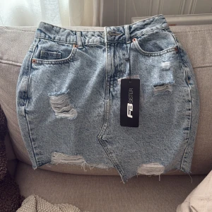 Blå jeanskjol med slitningar FB Sister - Säljer en snygg blå jeanskjol från FB Sister med coola slitningar och rå kant nertill. Kjolen har klassisk femficksdesign, bälteshällor och en asymmetrisk nederkant. Perfekt för dig som gillar en trendig och avslappnad stil. Aldrig använd endast testad.