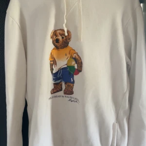 Vit hoodie Polo Bear Ralph Lauren M - Vit hoodie från Polo Ralph Lauren med ikoniskt Polo Bear-tryck framtill. Tröjan har huva med snörning och känguruficka. Mjuk insida och klassisk passform, perfekt för chill dagar. Materialet är bomull och polyester.