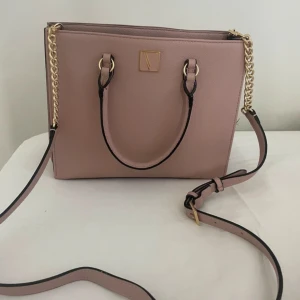 Rosa handväska från Victoria's Secret - Snygg ljusrosa handväska från Victoria's Secret med guldfärgade detaljer och kedja. Väskan har både handtag och axelrem, samt randigt foder och dragkedja upptill. Perfekt för dig som vill ha en stilren och trendig accessoar. Har nästan aldrig använt.