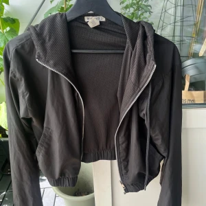 Svart croppad vindjacka Forever 21 XS - Svart croppad vindjacka från Forever 21 med huva och dragkedja framtill. Jackan har elastisk midja och ärmar, samt meshfoder på insidan för extra komfort. Perfekt för lager-på-lager och streetstyle.