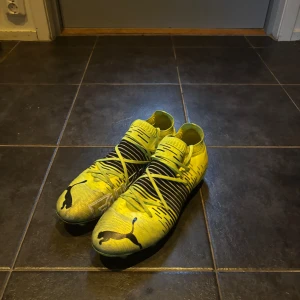 Gula fotbollsskor från Puma - Snygga gula fotbollsskor från Puma med svarta detaljer och coolt mönster. Skorna har snörning, rund tå och platt sula med dobbar för bra grepp på planen. Ovandelen är i syntetmaterial och designen är sportig och modern.