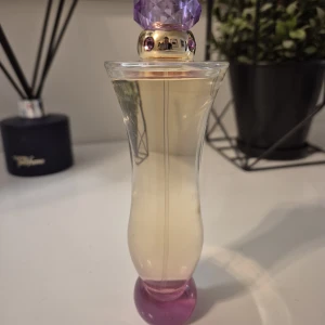 Versace  woamen  - Snygg parfymflaska med smal, böjd form och transparent glas. Flaskan har en lila kristallformad kork och guldiga detaljer med små lila stenar. Botten är rundad och rosa. Perfekt som present eller till din egen samling.