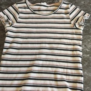 Randig ribbad topp från Lindex - Säljer en ribbad topp från Lindex med svarta, vita och ljusrosa ränder. T-shirten har rund halsringning och korta ärmar. Perfekt för dig som gillar en enkel men ändå snygg stil med lite retrovibbar.