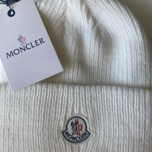 Vit ribbstickad mössa från Moncler med Tagg - Vit ribbstickad mössa från Moncler med klassisk uppvikt kant och broderad logotyp framtill. Snygg och enkel design som passar till det mesta. Tillverkad i mjukt material för extra komfort. 