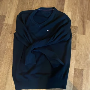 Svart v-ringad tröja från Tommy Hilfiger - Svart v-ringad tröja från Tommy Hilfiger med diskret logga broderad på bröstet. Klassisk och stilren modell med långa ärmar och ribbade muddar. Perfekt för dig som gillar enkel och snygg design.