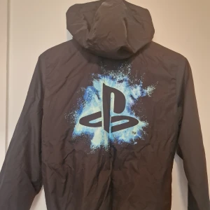 PlayStation vindjacka med huva - Svart vindjacka från PlayStation x HM med blått logotryck på ryggen och bröstet. Perfekt för sommaren. Mellan 10-12 år. 