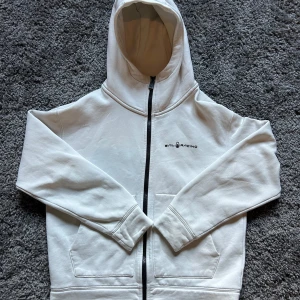 Vit hoodie från Sail Racing med dragkedja - Vit hoodie från Sail Racing med svart dragkedja och logga på bröstet. Tröjan har huva, två fickor fram och är tillverkad i mjuk bomullsmix. Perfekt för en chill och stilren look.