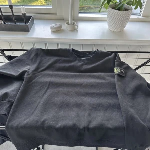 Svart sweatshirt från Stone Island - Svart sweatshirt från Stone Island med klassisk rund hals och ribbade muddar. Tröjan har den ikoniska Stone Island-loggan på vänster ärm. Tillverkad i mjuk bomull, perfekt för en clean och stilren look.