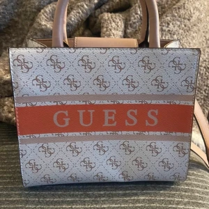 Beige och orange handväska från Guess - Snygg handväska från Guess i beige och orange med logomönster och bred orange rand med stor GUESS-text. Väskan har två rejäla handtag, magnetlås och flera fack inuti. Tillverkad i syntetmaterial och perfekt för dig som gillar statement-accessoarer.