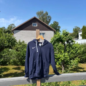 Mörkblå Nike hoodie med dragkedja - Snygg mörkblå hoodie från Nike med vit snörning och dragkedja framtill. Klassisk logga på bröstet och två stora fickor. Perfekt för chill dagar eller när du vill ha en sportig look. Mjuk insida och huva för extra komfort.