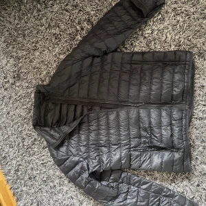 Svart dunjacka från J.Lindeberg - Säljer en svart dunjacka från J.Lindeberg med diskret logga på ryggen. Jackan är quiltad, har dragkedja framtill och två sidofickor med dragkedja. Sommarn kommer och går med vädret därför vore det nog skönt med en enkel jacka som den här! 