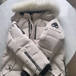 Beige dunjacka med pälskrage Moose Knuckles - Snygg beige dunjacka från Moose Knuckles med fluffig pälskrage, svarta detaljer och flera dragkedjor. Jackan har logga på ärmen och praktiska fickor. Perfekt för kalla dagar när du vill hålla stilen och värmen.