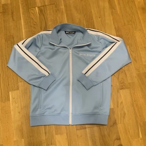 Ljusblå track jacket från Palm Angels - Palm Angels ljusblå track jacket med vita och svarta ränder längs ärmarna. Jackan har dragkedja framtill, hög krage och ribbade muddar. Tillverkad i 100% polyester för en sportig vibe. Perfekt för dig som gillar streetwear och snygga detaljer. men ganska fläckig
