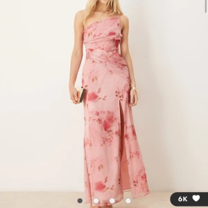 Rosa blommig långklänning med slits - Superfin långklänning i ljusrosa med blommigt mönster och en axel. Klänningen har en snygg slits framtill och omlott-detaljer i midjan. Tunn och flowy, perfekt för sommarens alla tillfällen. Axelbandslös på ena sidan för en cool look.