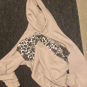 Beige hoodie med leopardmönster - Säljer en beige hoodie med huva och snyggt leopardmönstrat parti över bröstet. Tröjan har långa ärmar och en relaxed passform, perfekt för en chill och trendig look. Materialet känns mjukt och skönt mot huden.