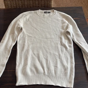 Beige stickad tröja från Selected Homme - Snygg vit stickad tröja från dressmanmed rund halsringning och lång ärm. Tröjan har en diskret ribbad struktur i muddar och nederkant, vilket ger en clean och stilren look. Perfekt att matcha med jeans eller chinos.