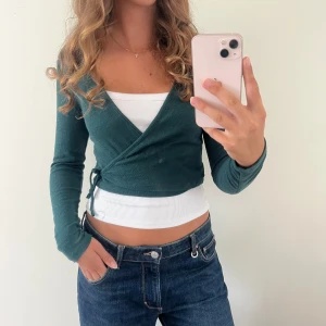 Mörkgrön omlott kofta med snörning - Snygg mörkgrön omlott kofta med snörning i sidan. Croppad modell med lång ärm och v-ringad hals. Perfekt att styla över ett linne 