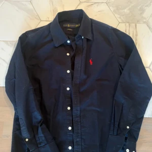 Mörkblå skjorta från Ralph Lauren - Klassisk mörkblå skjorta från Ralph Lauren med vit knappstängning och röd broderad logga på bröstet. Skjortan har lång ärm och stilren krage, perfekt för en snygg och enkel look. Tillverkad i bomull för en skön känsla.