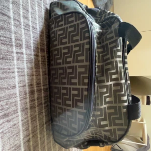 Mörkgrön Fendi resväska - Säljer en snygg Fendi resväska i svart och grått med grafiskt mönster. Väskan har dragkedja upptill, bärhandtag i nylon och rymligt huvudfack. Perfekt för weekendresor eller gymmet. Stilren design som sticker ut.
