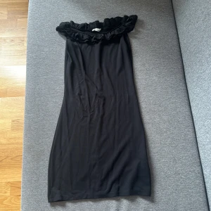 Sommarklänning - svart - Svart axelbandslös klänning med bred volang upptill. Snygg och enkel modell som sitter offshoulder och har en rak, något figurnära passform.