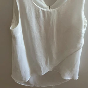 Vit omlott-linnetopp från Bershka - Säljer en vit ärmlös linne-topp med omlott-detalj framtill och v-ringning både fram och bak. Toppen har en lätt och luftig känsla, perfekt för varma dagar. Stängs med en liten knapp i nacken och har en snygg asymmetrisk nederkant