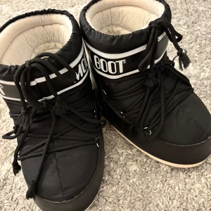 Svarta Moon Boots - Moon Boots i svart med vit logga och detaljer. Fodrade och riktigt varma, perfekta för vintern. Klassisk rund form, snörning framtill och robust sula med grepp. Yttermaterial i syntet och insida i mjukt tyg. En ikonisk vinterboot som sticker ut i snön.