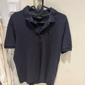 Svart pikétröja från BOSS - Klassisk svart pikétröja från BOSS med broderad logga på bröstet. Tröjan har krage, knappar framtill och korta ärmar,använd men inget slitage,storlek M