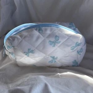 Quiltad vit necessär med blå rosetter - Supergullig quiltad necessär i vitt med ljusblå rosett-mönster och matchande blå dragkedja. Perfekt för smink eller småprylar, mjuk och lätt att ta med sig. Söt design som sticker ut i väskan eller på hyllan.