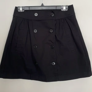 Svart kortkjol med knappar från Esprit - Svart kortkjol från Esprit med dubbelknäppning framtill och två fickor. Kjolen har en klassisk A-linjeform och är gjord i bomull, vilket gör den både bekväm och lätt att matcha. Perfekt för dig som gillar stilrena plagg.